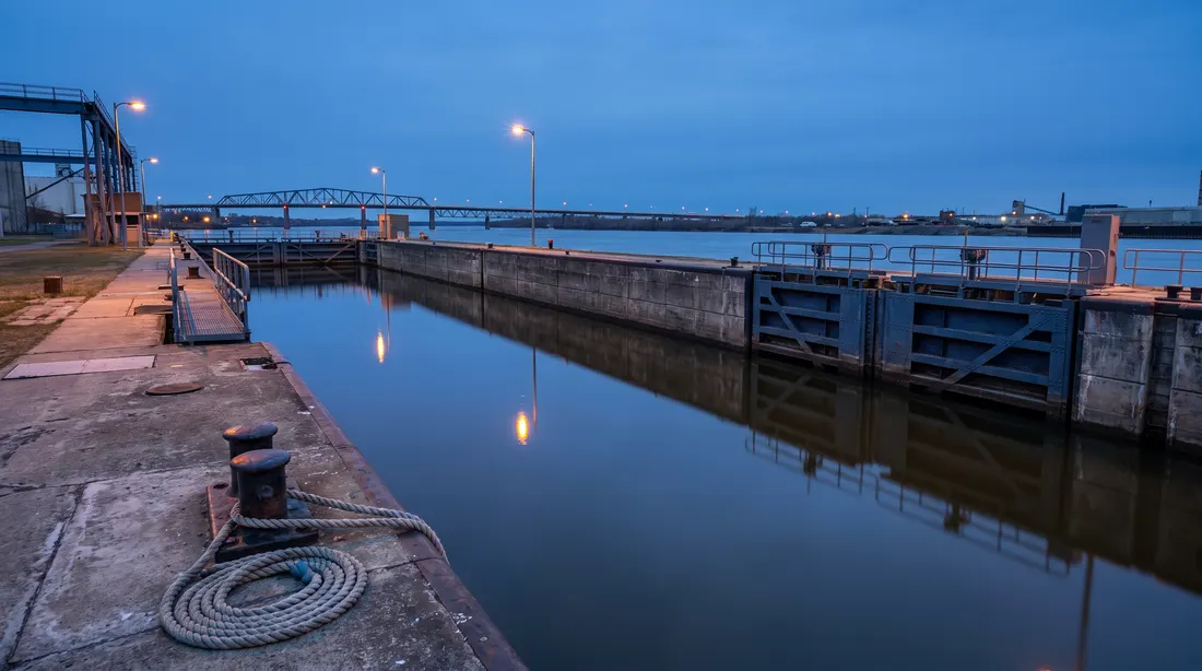 Generated scene: a canal lock at blue hour, two closed steel gates at the end of a long water-filled chamber, a steel truss bridge in the distance; not a photograph of the casino.